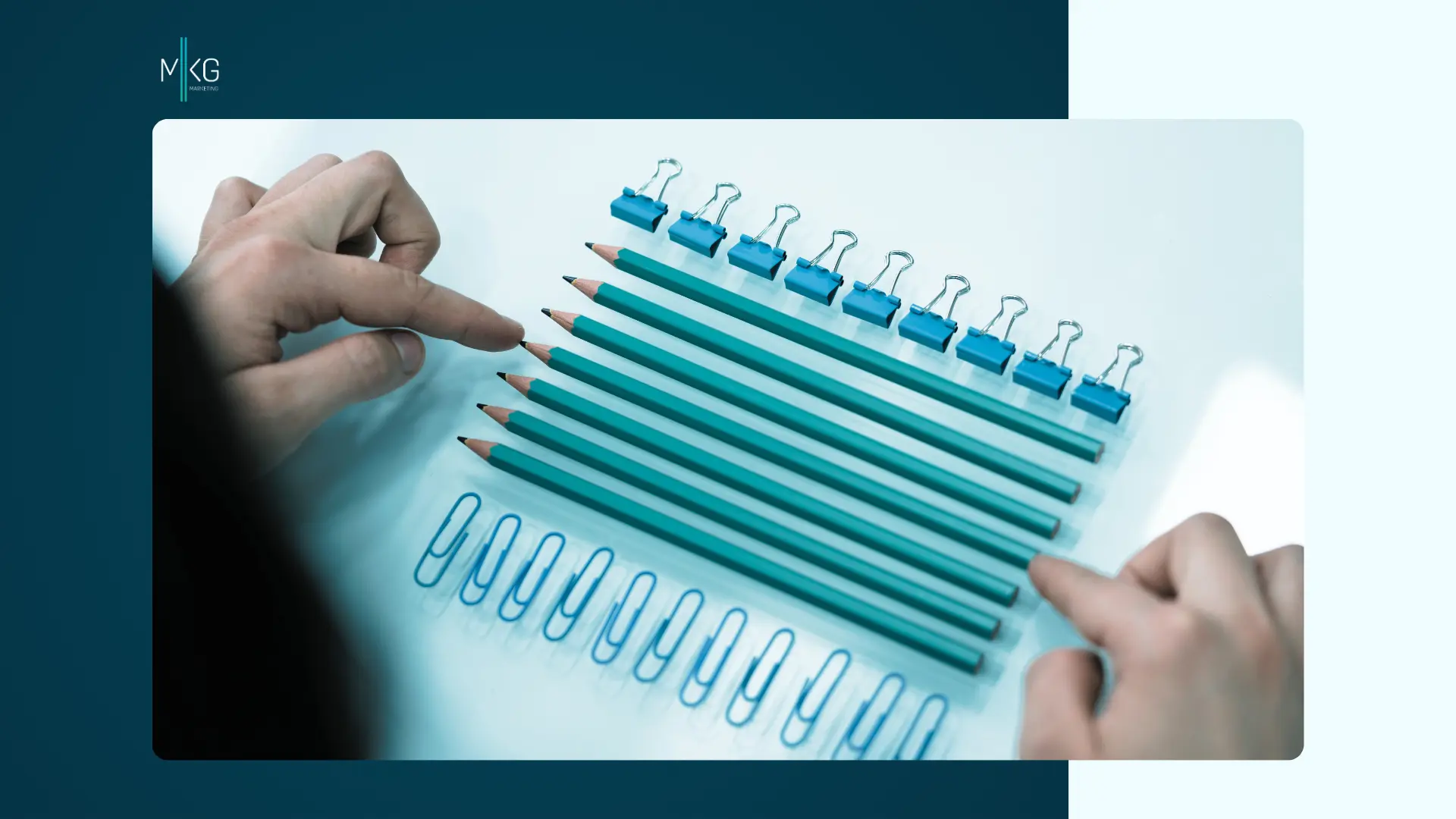A high-angle view of two hands carefully aligning teal pencils arranged neatly between rows of blue binder clips and teal paper clips on a white surface, with an "MKG Marketing" logo in the top-left corner.